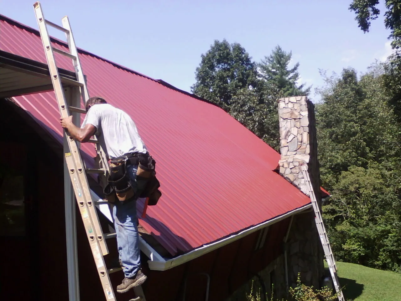 Roofer on a ladder beside a red metal roof and stone chimney — Soffit & Fascia Repair by Roofing Brothers in Elverson